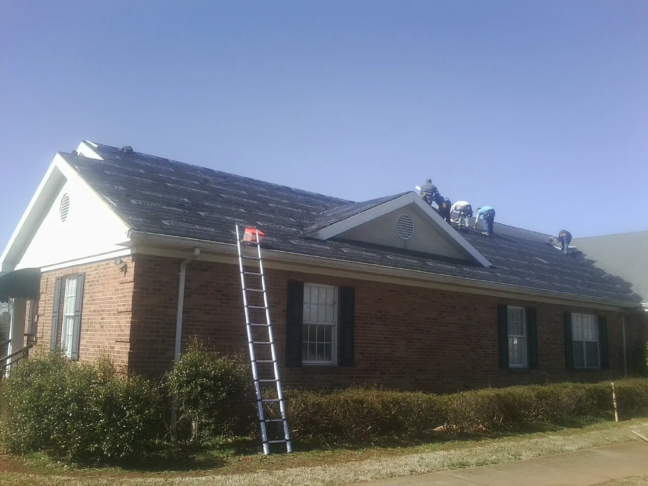 Crew tearing shingles off a brick ranch roof with a ladder set — Emergency Roof Repair work by our crew in Chatham